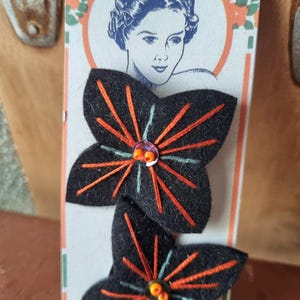 Puede incluir: Dos horquillas de fieltro negro con forma de flor, con costuras naranjas y turquesas y una cuenta central. Las horquillas se muestran en una tarjeta vintage con una ilustración azul del rostro de una mujer.