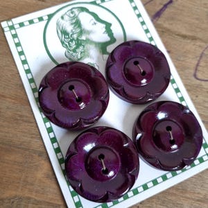 Può includere: Quattro bottoni viola scuro a forma di fiore con finitura lucida. Ogni bottone ha un design a due fori. I bottoni sono disposti su una carta bianca con un bordo verde e un'illustrazione vintage del profilo di una donna.