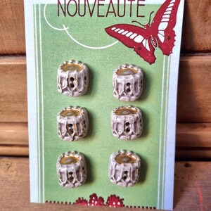 May include: Six white ceramic buttons with gold accents on a green card with the word "NOUVEAUTÉ" and a red and white butterfly.