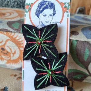Puede incluir: Dos horquillas para el pelo con forma de flor de fieltro negro, con detalles bordados en verde y rojo y una cuenta central. Las horquillas están en una tarjeta vintage con un retrato de mujer y el texto "MADE IN FRANCE".