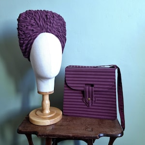 Peut inclure: Un chapeau de style turban de couleur prune est posé sur une tête de mannequin blanche, exposé sur un support en bois. À côté, un sac à bandoulière à rayures prune assorti avec une fermeture à rabat et une longue sangle.