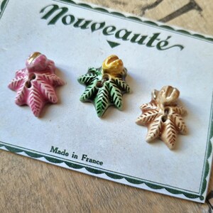 Peut inclure: Trois boutons en céramique avec un motif de feuilles. Les boutons sont rose, vert et beige avec des accents dorés. Les boutons sont sur une carte blanche avec le texte "Nouveauté" et "Fabriqué en France".