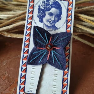 Puede incluir: Una horquilla de fieltro azul marino con forma de flor con costuras turquesas y rosas, y un centro de cuentas doradas. La horquilla está unida a una tarjeta vintage con un borde geométrico azul y naranja y un retrato de mujer.