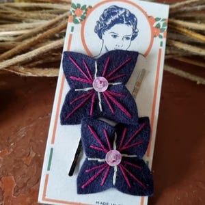 Puede incluir: Dos horquillas para el pelo de fieltro azul marino con forma de mariposa, con detalles bordados en rosa y un botón rosa en el centro. Las horquillas están en una tarjeta de estilo vintage con un retrato de mujer y el texto "Made in France".