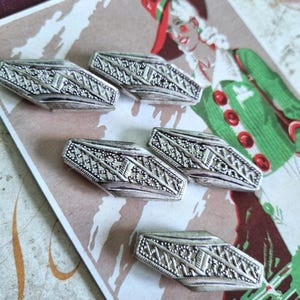 May include: Six silver-toned metal buttons with intricate geometric designs. The buttons are arranged on a vintage illustration featuring a person in a green coat and red hat. The buttons have a rectangular shape with detailed patterns.