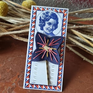 Puede incluir: Una pinza para el pelo de estilo vintage con una flor de fieltro azul marino con costuras rosas y amarillas. La pinza está unida a una tarjeta con un retrato de mujer y un borde geométrico azul, naranja y blanco.