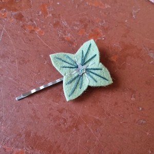 Puede incluir: Una horquilla de fieltro verde claro con forma de trébol de cuatro hojas. El trébol tiene venas bordadas en verde oscuro y una pequeña joya plateada en el centro. La horquilla está unida a una horquilla plateada.