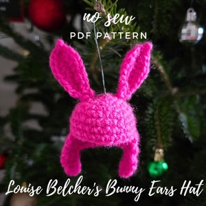 Op de afbeelding: Roze gebreide bunny oren muts met de tekst "no sew PDF pattern" en "Louise Belcher's Bunny Ears Hat" hangend aan een kerstboom.