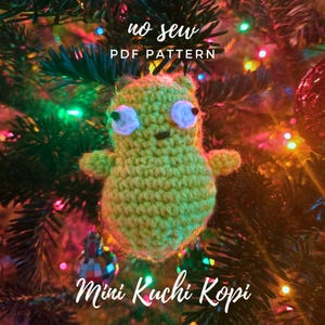 Op de afbeelding: Een groen gehaakt amigurumi-speelgoed in de vorm van een personage uit de show "Kuchi Kopi". Het speelgoed hangt aan een kerstboom met kleurrijke lampjes. De tekst "no sew PDF pattern" en "Mini Kuchi Kopi" is zichtbaar in de afbeelding.