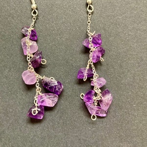 Könnte beinhalten: Ohrringe mit Amethyst-Edelsteinen. Jeder Ohrring hat einen silberfarbenen Haken und eine Kette, an der Büschel aus lila und lavendelfarbenen Amethyststeinen hängen.