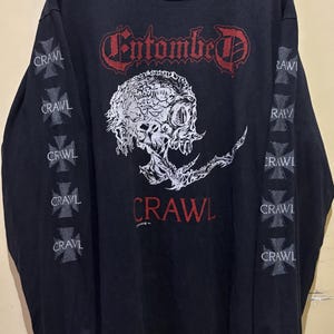 Puede incluir: Camiseta negra de manga larga con el nombre de la banda "Entombed" en letras góticas rojas y un gráfico de calavera. Las mangas muestran la palabra "Crawl" y diseños de cruces. Una camiseta vintage de banda.