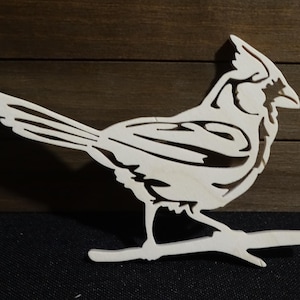 May include: A white wooden cutout of a cardinal bird perched on a branch. The bird is detailed with intricate lines and a crest on its head.
