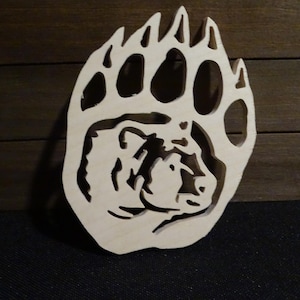 May include: A wooden bear paw print with a bear face carved into the center. The paw print is a light brown color and the bear face is a darker brown color.