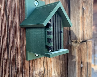May include: A green, 3D printed insect house with a black slatted entrance. The house is mounted on a wooden surface.