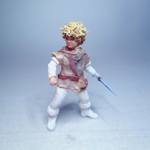 May include: A small, hand-painted figurine of a young person with blond hair wearing a brown tunic and white pants. The figurine is holding a small, blue sword.