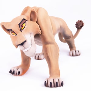 May include: A brown lioness figurine with a fierce expression, bright orange eyes, and a black nose. The figurine is posed in a crouching position with its paws extended forward.