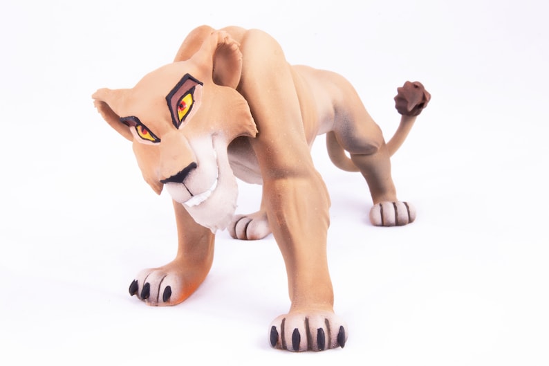 May include: A brown lioness figurine with a fierce expression, bright orange eyes, and a black nose. The figurine is posed in a crouching position with its paws extended forward.