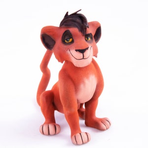 May include: A red and brown lion cub figurine with black hair and yellow eyes. The figurine is sitting on a white surface.