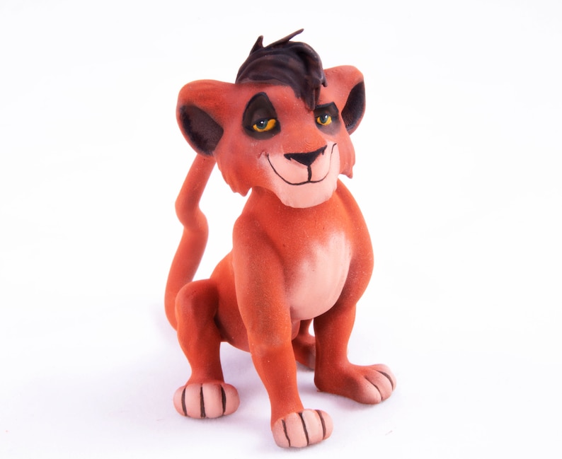 May include: A red and brown lion cub figurine with black hair and yellow eyes. The figurine is sitting on a white surface.