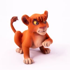 May include: A small, orange lion cub figurine with a grumpy expression. The figurine is made of a textured material and has a detailed face with yellow eyes.