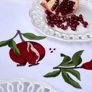Puede incluir: Un mantel blanco con bordados detallados de granadas rojas y hojas verdes. La tela está decorada con múltiples motivos de granadas y pequeños puntos rojos. Se observan dos platos de cerámica blanca con bordes de celosía.