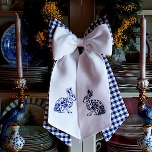 以下が含まれることがあります： 白地に青と白のギンガムチェックのトリムが付いた白いリボンが、青と白の磁器の棚のある白いキャビネットにかけられています。リボンには、青い刺繍のウサギが2匹飾られています。