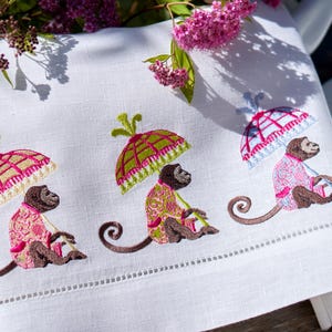 Puede incluir: Toalla de lino blanco bordada con tres monos, cada uno sosteniendo un paraguas. Los monos visten ropa estampada rosa y verde. Los paraguas son rosas, verdes y azules. La toalla tiene un borde cosido decorativo.