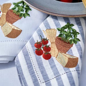 Op de afbeelding: Geborduurde servetten met Italiaanse voedselmotieven. De wit en blauw gestreepte servetten tonen borduursels van tomaten, basilicum, pasta en kaas. De servetten zijn gevouwen in de buurt van een bord en spaghetti.