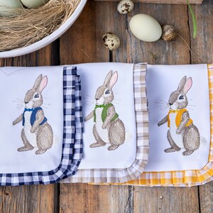 May include: Three white tea towels with embroidered brown rabbits wearing different colored vests. The towels have gingham borders in blue, beige, and yellow.