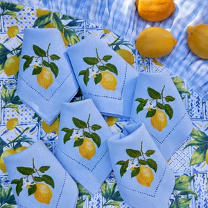 May include: Six light blue linen napkins with embroidered lemon designs on a white and blue patterned tablecloth. The napkins are folded and arranged in a circle.