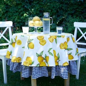 Pu&ograve; includere: Una tovaglia rotonda bianca con stampa di limone e un bordo a quadri blu e bianco. La tovaglia &egrave; posta su un tavolo con due sedie bianche e un'ampolla di limonata.