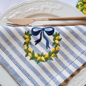 May include: A blue and white striped linen napkin with an embroidered lemon wreath and a blue bow. The napkin is folded and placed on a white plate.