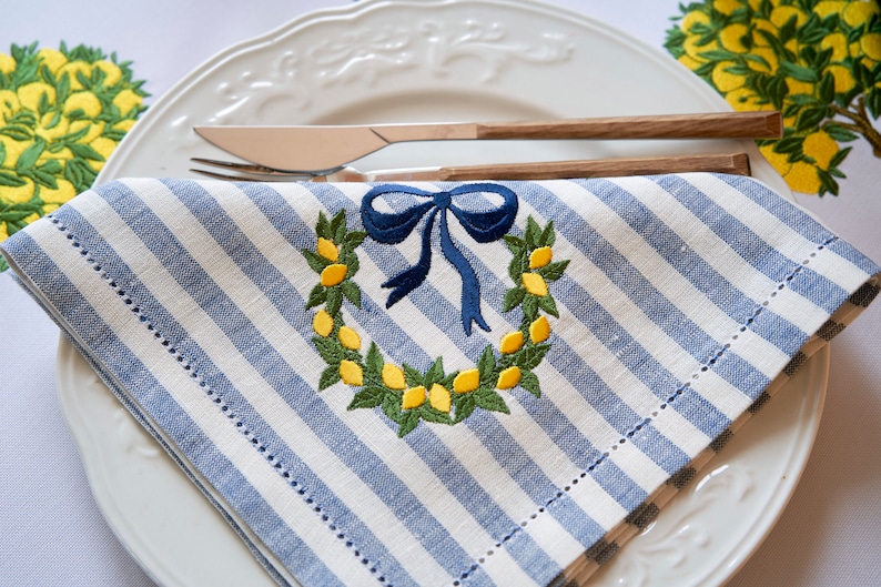May include: A blue and white striped linen napkin with an embroidered lemon wreath and a blue bow. The napkin is folded and placed on a white plate.
