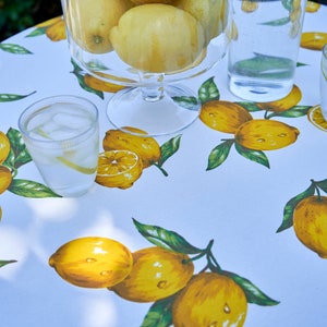 Pu&ograve; includere: Una tovaglia bianca con stampa di limoni. Una ciotola di vetro piena di limoni si trova sul tavolo, insieme a una caraffa di limonata e due bicchieri di acqua ghiacciata.
