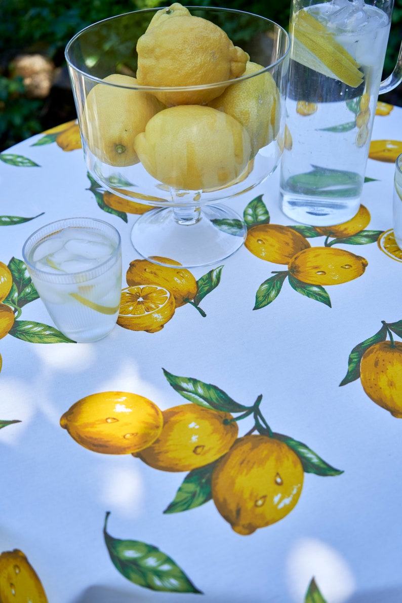 Pu&ograve; includere: Una tovaglia bianca con stampa di limoni. Una ciotola di vetro piena di limoni si trova sul tavolo, insieme a una caraffa di limonata e due bicchieri di acqua ghiacciata.
