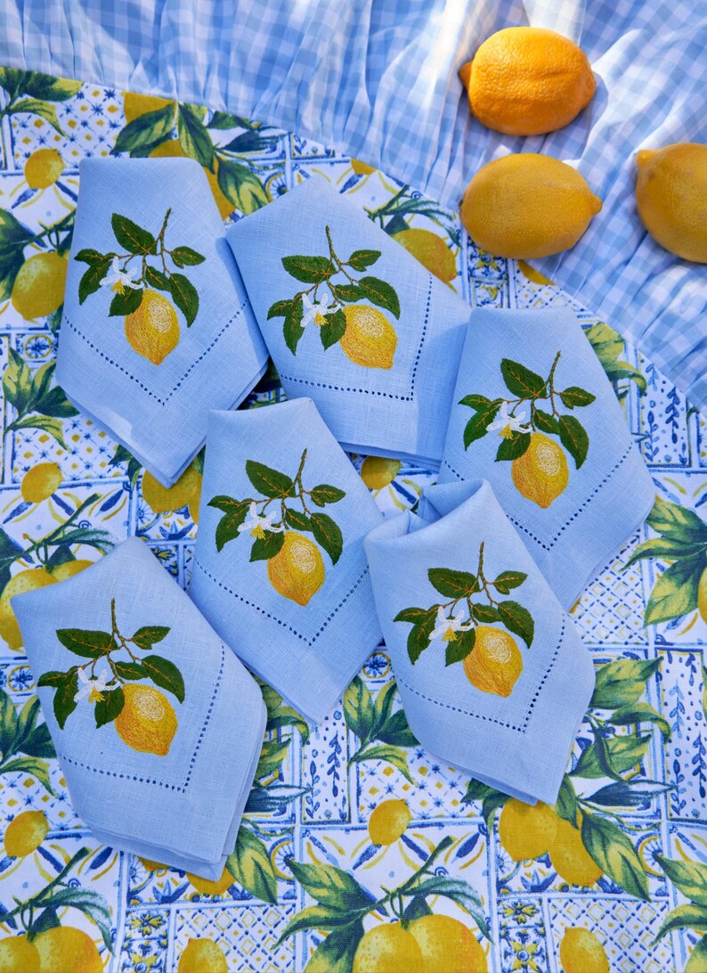 May include: Six light blue linen napkins with embroidered lemon designs on a white and blue patterned tablecloth. The napkins are folded and arranged in a circle.