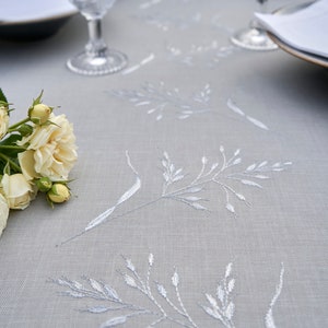 May include: A gray linen tablecloth with white embroidered floral designs. A bouquet of white roses is placed on the tablecloth.