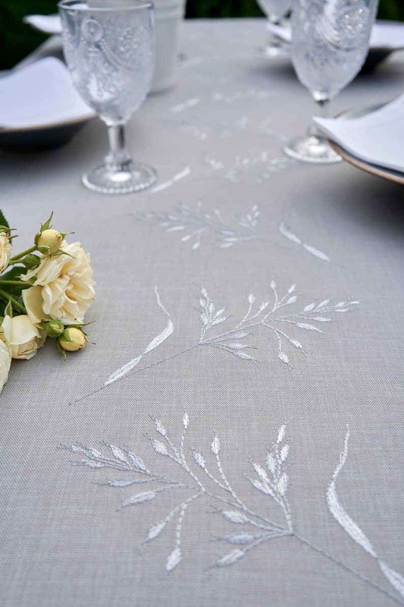 May include: A gray linen tablecloth with white embroidered floral designs. A bouquet of white roses is placed on the tablecloth.
