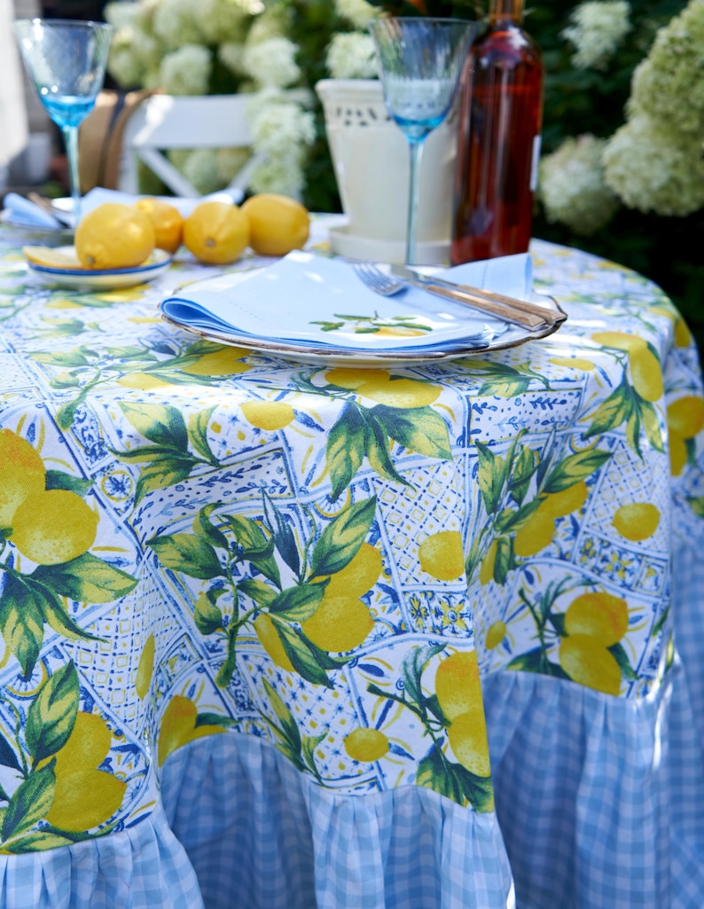 May include: A blue and white tablecloth with a lemon print and a gingham border. The tablecloth is set with a white plate, a blue napkin, and silverware.