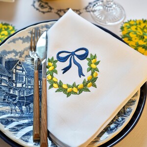 May include: A white linen napkin with a blue ribbon and a wreath of green leaves and yellow lemons embroidered on it. The napkin is folded and placed on a blue and white patterned plate with a fork and knife.