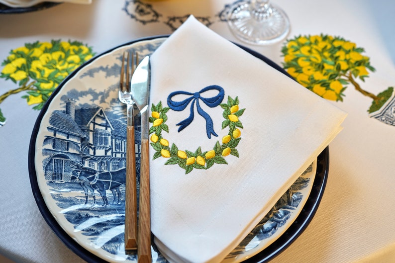 May include: A white linen napkin with a blue ribbon and a wreath of green leaves and yellow lemons embroidered on it. The napkin is folded and placed on a blue and white patterned plate with a fork and knife.