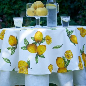 Pu&ograve; includere: Tovaglia rotonda bianca con stampa di limoni. La tovaglia ha un bordo arricciato ed &egrave; sovrapposta a una tovaglia bianca liscia. Ci sono due bicchieri d'acqua e una caraffa di limonata sul tavolo.