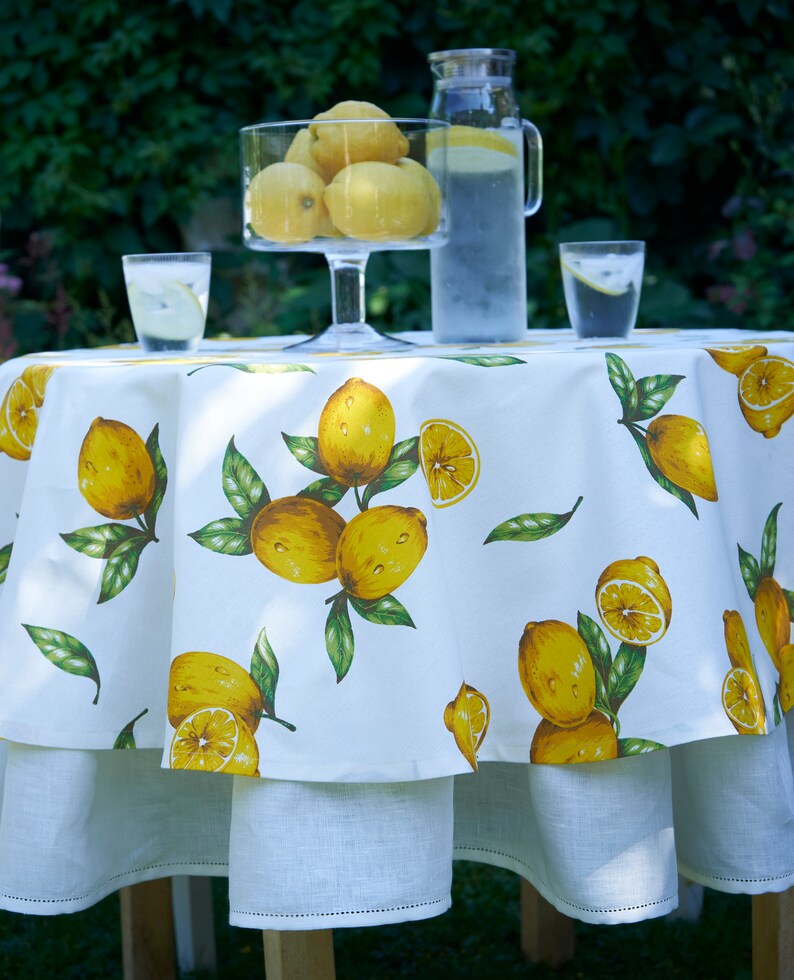 Pu&ograve; includere: Tovaglia rotonda bianca con stampa di limoni. La tovaglia ha un bordo arricciato ed &egrave; sovrapposta a una tovaglia bianca liscia. Ci sono due bicchieri d'acqua e una caraffa di limonata sul tavolo.