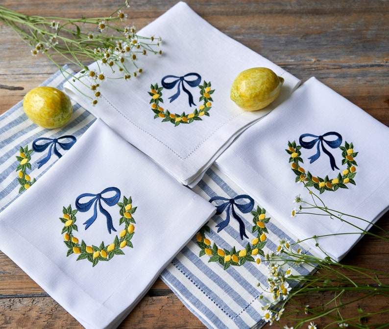May include: Four white linen napkins with blue and yellow embroidered lemon wreaths and blue bows. The napkins are arranged on a blue and white striped surface with lemons and white flowers.