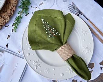 Può includere: Un tovagliolo di lino verde con un ricamo a forma di albero di Natale è piegato e posizionato su un piatto bianco con un motivo a fiocco di neve. Il tovagliolo è legato con un anello per tovaglioli in juta marrone.