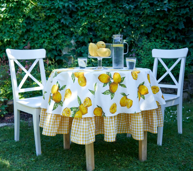 Pu&ograve; includere: Tavolo bianco con una tovaglia a quadri gialla e bianca con stampa di limoni. Due sedie bianche sono posizionate su entrambi i lati del tavolo. Una caraffa di vetro d'acqua e due bicchieri sono sul tavolo.