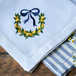 May include: White linen napkin with a blue ribbon and a wreath of yellow lemons embroidered on it. The napkin is folded and placed on top of a blue and white striped napkin.