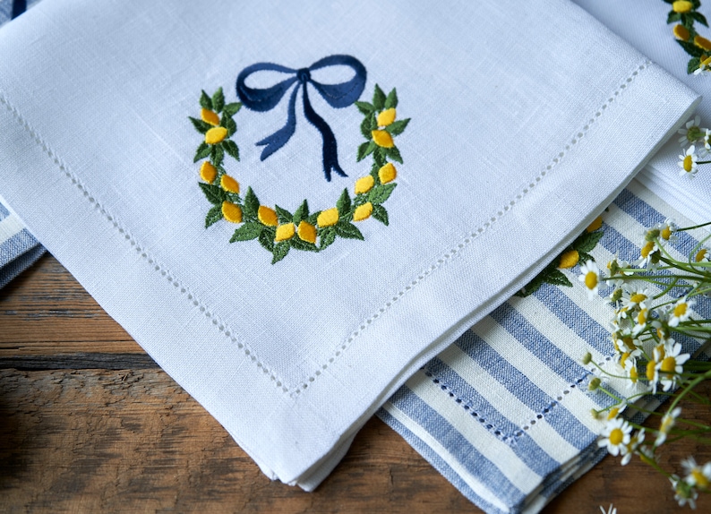 May include: White linen napkin with a blue ribbon and a wreath of yellow lemons embroidered on it. The napkin is folded and placed on top of a blue and white striped napkin.