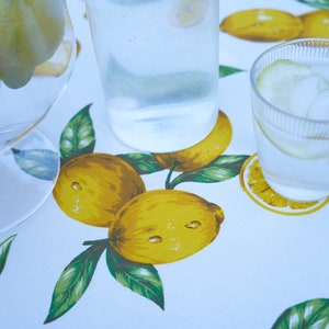 Pu&ograve; includere: Una tovaglia bianca con stampa di limone presenta una caraffa di vetro trasparente piena d'acqua e un bicchiere d'acqua con ghiaccio e una fetta di limone.