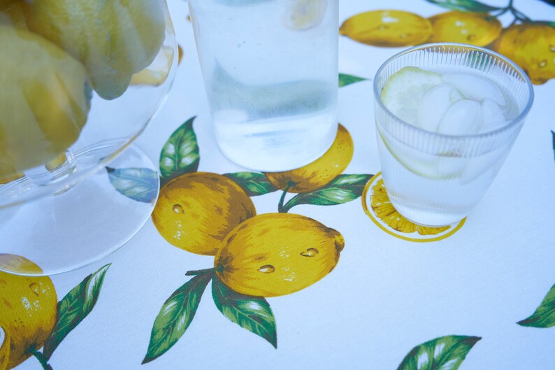 Pu&ograve; includere: Una tovaglia bianca con stampa di limone presenta una caraffa di vetro trasparente piena d'acqua e un bicchiere d'acqua con ghiaccio e una fetta di limone.